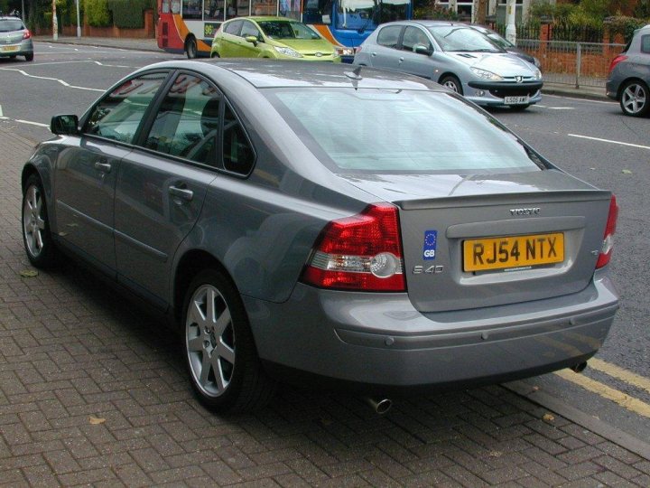 Back to the Volvo fold - Page 1 - Volvo - PistonHeads - The image shows a side view of a silver car parked on a brick paved side of a street. The car has a yellow license plate with black lettering, and there are other vehicles and cars in the background. The street appears to be in an urban area with traffic control lines and fencing visible. A globe is placed securely with its metal arm in the trunk.