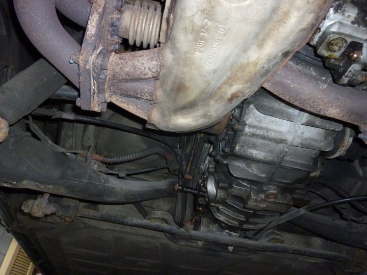 Pistonheads - This image depicts a view of the undercarriage of a vehicle, focusing on the front of the engine bay. The motor housing appears to be made of metal and has visible dirt, rust, and residual grease. A black flexible hose is connected to the engine block, and there are various mechanical components visible, including a pipe and a series of wires. The overall condition suggests it might be a repair or a vehicle that is not in active use. The angle of the photo provides a visible path of the motor housing leading down to the unseen parts of the vehicle.