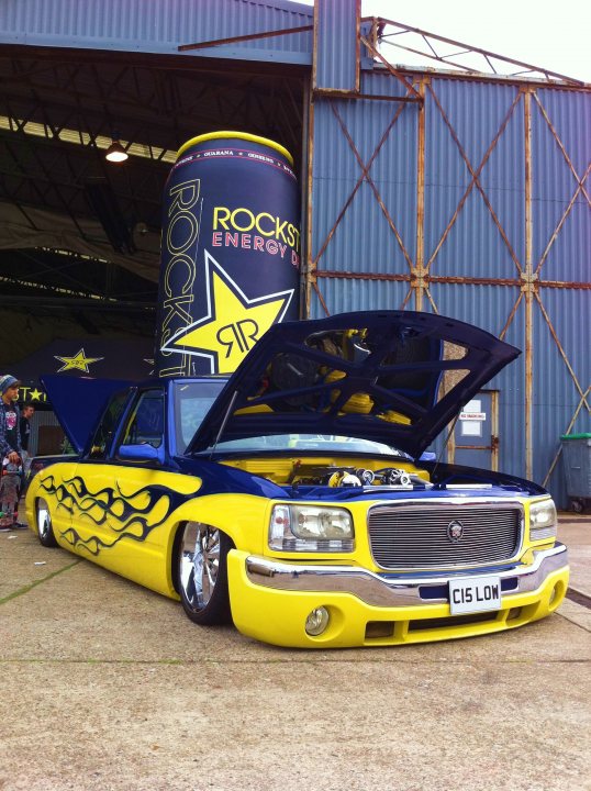 Pistonheads Chevy Truck - In the image, a modified yellow luxury car, possibly a customized Hummer or Range Rover, is parked in front of a metal building. The vehicle's design is notable for its stretched body and blue graphics, with flames painted on the hood. A massive tall can, reminiscent of the Monster Energy brand, is placed next to the car, creating an intriguing juxtaposition between the luxurious vehicle and the urban energy drink culture. The setting appears to be an outdoor parking area, indicated by the presence of a person in the background, adding a sense of scale to the image.