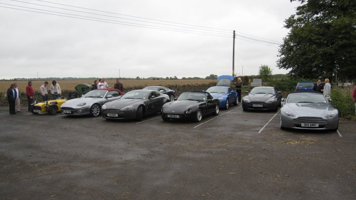 Pistonheads - The image captures an impressive lineup of luxury cars parked in a row in an outdoor setting. These cars are lined up by their models, creating a visual comparison of their sleek designs. The cars, predominantly black and gray, are parked in a gravel lot, and the people standing nearby suggest that this might be a display at a car show or some sort of event. The setting implies a relaxed atmosphere, possibly in a countryside location, with a vast open sky formed by the joining of clouds.