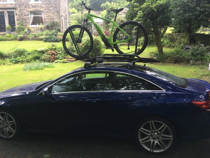 Ebike with battery integral to frame - roofrack options - Page 1 - Pedal Powered - PistonHeads - In the image, a blue sports car is parked on a grass lawn. Resting on the roof of the car is a green mountain bike. The car is facing slightly to the right of the frame, giving the impression that it is ready to drive off. The scene is bathed in natural light, suggesting it was taken during the day. The bike's position on top of the car indicates an active lifestyle and perhaps someone who enjoys mountain biking. The car itself appears to be well-maintained and sleek, reflecting the owner's taste.