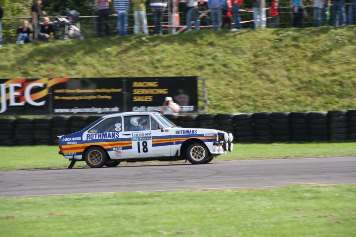 Day Coombe Castle Rally September Pistonheads Saturday - In the image, a blue and white race car from the Rothmans team is seen driving on a racing track. The car is coming towards the camera, with people visible in the background. The track fence indicates that this could be a race track. The car is numbered 18 and is equipped with sponsor decals.