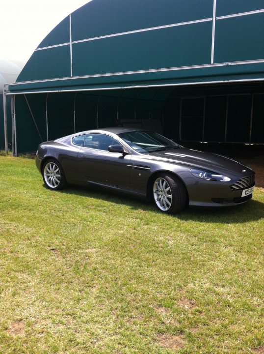 Pistonheads - This image features a luxurious, dark-colored Aston Martin sports car parked in a garage area. The car is sleek and elegant, suggesting high performance capability. It's positioned at a three-quarter angle, highlighting the profile and alloy wheels. Shadows cast on the verdant lawn indicate that the photo was taken during the day under natural light.