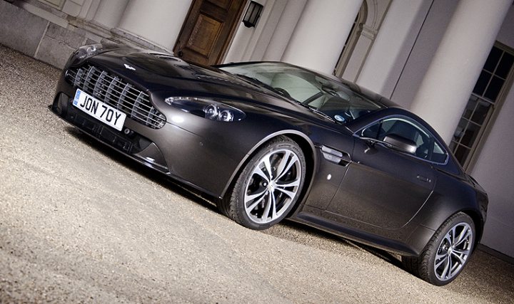 My V12V photographed by a talented photographer (not me) - Page 1 - Aston Martin - PistonHeads - The image showcases a sleek black Aston Martin Vanquish sports car, which is parked on a gravel surface. The car has a license plate on the front and is positioned at an angle that slightly obscures the front wheel. In the background, there's a columned structure that adds a touch of classical architecture to the scene. The lighting suggests it might be evening or in a dimly lit setting, which further emphasizes the car's design and color.