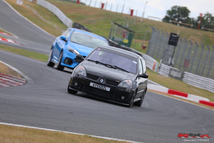 Clio 182 Track Toy - Page 2 - Readers' Cars - PistonHeads - The image captures a thrilling moment on a curvy racing track. Two cars are in an intense competition. A blue car is in the lead, skillfully navigating the left side of the track, while a black car is close behind, racing on the right side of the track. They both appear to be Renault cars, suggesting a match between two Renault vehicles. In the background, a flag is visible, likely signaling the ongoing race.