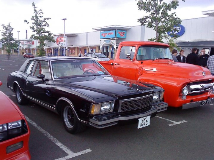 Pistonheads Edinburgh Cruisemeet - The image depicts a parking lot where two vintage cars are parked side by side. The car on the left appears to be a black classic with a bright orange one on the right. The cars are adjacent to a building, which adds a commercial backdrop to the scene. There are a few people visible in the parking lot, suggesting that this might be a popular spot, possibly frequented by car enthusiasts. The cars are parked neatly between white lines, adhering to a well-maintained parking lot.