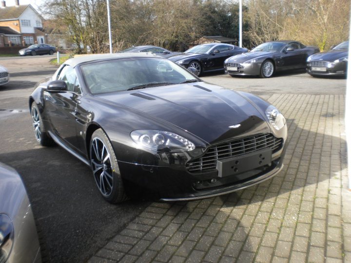 Pistonheads - The image shows a parking lot with several sports cars parked on a brick pavement. The car in focus in the foreground is a sleek, dark-colored sports car, possibly an Audi, due to its distinctive front grille. The car in the background on the right is an Aston Martin, identifiable by its unique distinctive grille and wing, which is typically associated with this luxury vehicle brand. The atmosphere of the image suggests a showroom or dealer environment.