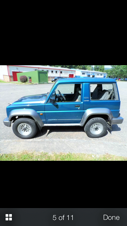 A blue truck parked in a parking lot - Pistonheads - This is a photograph of a blue SUV parked on a concrete surface. The vehicle appears to be a four-door model with visible rear-view mirrors and a spare tire mounted on the side of the body. There are no visible text, license plates, or distinctive insignia in the image. The setting seems to be an outdoor, possibly urban, area as indicated by other buildings and structures in the background.