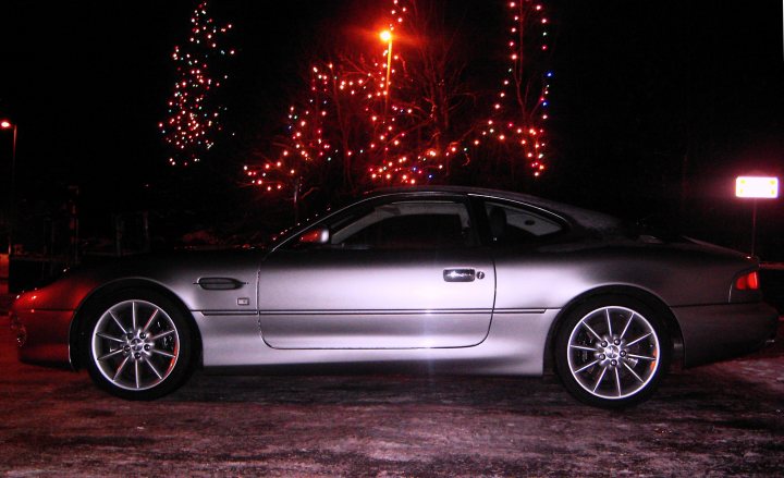 The *AM in the snow* thread :) - Page 2 - Aston Martin - PistonHeads - The image shows a sleek, dark-colored luxury car parked on a street at night. The vehicle's glossy finish reflects the ambient light, which is accentuated by festive Christmas lights adorning the surrounding trees and a street sign with a partially visible message. The street is not crowded, and the overall atmosphere is calm and serene, highlighting the elegance of the car.