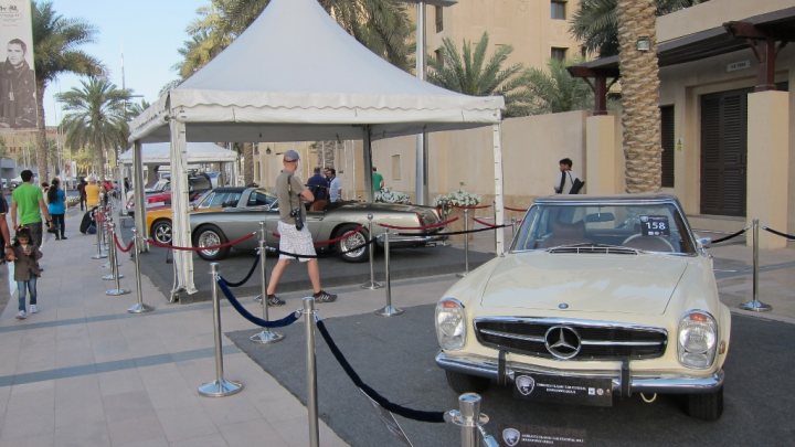 Anybody up for a trip to the emirates classic car festival  - Page 1 - Middle East - PistonHeads - The image showcases an outdoor exhibit featuring vintage cars, including a prominent display of a Mercedes Benz sports car. The cars are placed behind metal barricades and velvet ropes, indicating a view-only exhibit. Several individuals are present in the scene, with two people near the exhibit and another individual more distantly. The setting suggests a public event or exhibition, possibly held on a sidewalk in front of a building. The display of the cars against the backdrop of the building implies an organized and formal event.