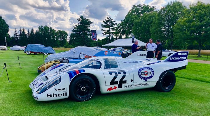 The Porsche's Big 70th Anniversary adventure... - Page 2 - General Gassing - PistonHeads - The image showcases a vintage race car, the unique Shell Martini Racing Team livery, number 27, white with blue and red stripes, prominently displayed. The car is parked on a lush grass field, surrounded by other vintage race cars. Men in polo shirts are interacting near the car, perhaps discussing the vehicle or related matters. Signage, possibly identifying the Porsche teams or other elements of the event, can also be observed in the background.