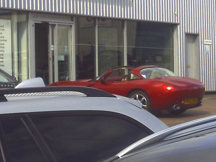Pistonheads - The image captures a lively street scene featuring a vibrant red sports car parked next to what appears to be a silver car. The sports car is prominently positioned in the view, drawing attention with its sleek design and bright color. The silver car, partially visible, is parked right next to it, creating a striking contrast between the two vehicles. In the background, there's a building adorned with signs, suggesting a commercial area. A person can be seen in the distance, adding a touch of life to the scene.