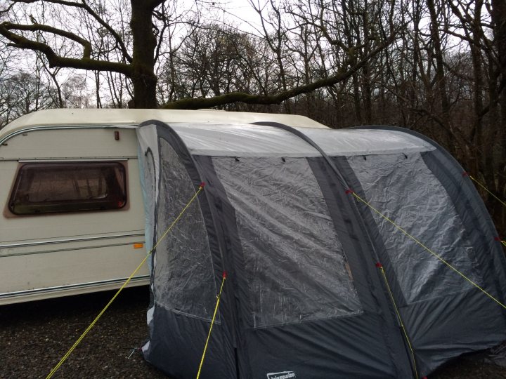 Awning Problems - Page 1 - Tents, Caravans & Motorhomes - PistonHeads - This image shows a caravan camping scene featuring a Trojan brand awning tent. The tent is silver and stands next to a cream-colored caravan on a gravel surface. Tall, bare trees surround the area, creating a serene outdoor setting. The tent is secured to the caravan with yellow ropes, indicating that it is being used. The overall scene suggests an outdoor recreational activity or camping trip.