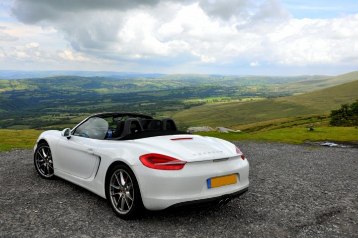 New Arrival! White Boxster S with PSE - Page 1 - Porsche General - PistonHeads - The image captures a striking scene of a white Porsche sports car parked on a gravel surface. The car is positioned in the bottom right corner of the frame, with a panoramic view of a lush valley and mountains in the background. The sky is dotted with clouds, suggesting a partly cloudy day. The setting appears serene and the car is the main focal point in the foreground. The overall composition of the image highlights the contrast between the rugged terrain and the sleek luxury of the sports car.