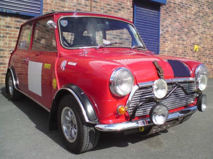 Breakfast Easter Malton Big Pistonheads Sunday April Yorkshire - The image shows a classic red Mini Cooper parked beside a brick wall. The car is adorned with various stickers and a distinctive smuggler's stripe. It features a unique chrome bumper and has a classic round front headlight configuration. There's no visible text on the hood or the windshield of the car. The Mini Cooper has a straightforward, all-body color scheme without any patterns or designs in the photo.