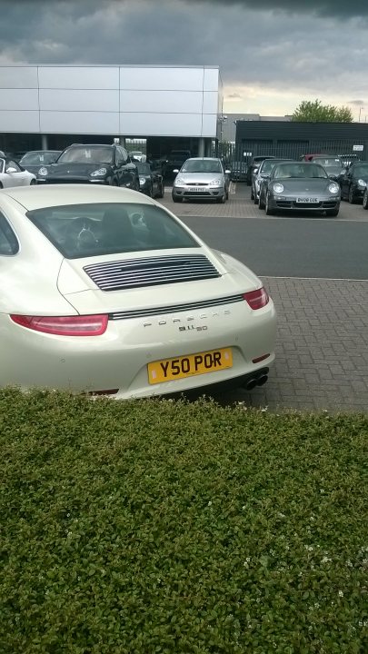 Real Good Number Plates : Vol 4 - Page 348 - General Gassing - PistonHeads - The image shows a scene in a parking lot. A white Porsche is the main focus, parked adjacent to a grassy area dotted with white flowers. Just behind the Porsche, there is a row of other vehicles, including a blue car, partially obscured by the vibrant greenery. The backdrop of the scene is a cloudy sky, with buildings visible in the background. The overall atmosphere suggests an early evening setting.