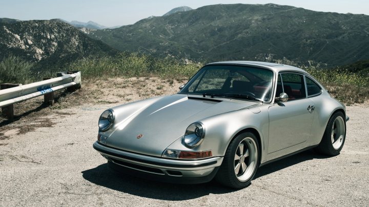 The Best Looking 911? - Page 1 - 911/Carrera GT - PistonHeads - The image features a silver Porsche speedster positioned in the center, parked on a gravel surface with a mountainous backdrop. The car, with its sleek design and white decals, is angled to the left, facing away from the grand landscape. The mountain range is lush with greenery, and there's a guard rail in the foreground that contrasts with the rugged terrain behind. This scene captures a sense of adventure and the allure of travel.