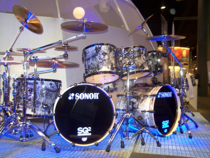 A group of people standing in front of a clock - The image showcases a large electronic drum set on display. The drums are dark with silver accents, displaying the Sonor brand, and appear to be in the year make model, as indicated by the SQ2 on a drum. There is a circular metallic pedal in the foreground and a cymbal attached to the stand. An additional electronic pad is in the background. The drum set is illuminated with various blue lights against a ceiling with glass panels, giving the impression that the set is behind a glass displayable.