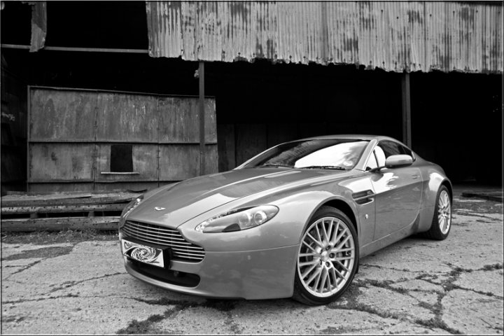 Thread Aston Pistonheads - In the middle of a street with cracked asphalt, a sleek, green Aston Martin DB9 coupe is parked against an old wooden fence. The car, which is small and luxurious, is well-lit by the sun, highlighting its shiny surfaces. The fence shows signs of wear and age, contrasting with the timeless beauty of the sports car. The surrounding structures suggest a quiet neighborhood with minimal activity.