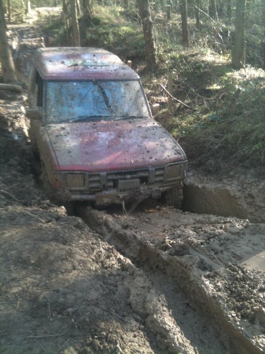 Pistonheads - The image depicts a beaten-up subaru car that seems to have been caught in a rough off-road terrain, now part of a thick mud track. The car appears heavily used with visible mud coating its body and the surrounding area. Its front end is almost submerged in the mud, and only a part of its body can be seen above the muddy path. There are remnants of various dense plants indicative of a forested area, and the clearing through which the mud track passes is visibly obstructed by thick vegetation. The sunlight filtering through the trees casts shadows over the car and the nearby area, creating a sense of depth and light in the image.