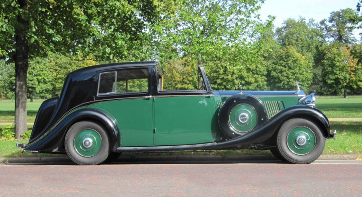 Prive Concours Delegance Pistonheads Salon - The image depicts a classic four-door green sedan parked on the side of a road. The car features a vintage design with a ribbed front grille, twin round headlights, and a prominent nose piece. Its windows are made of glass, and there is a visible chrome hood ornament. The car is parked in what appears to be a suburban or park area, as indicated by the lush green trees and a park-like atmosphere in the background. The vehicle is facing towards the right side of the image, and there is a clear view of its side profile.