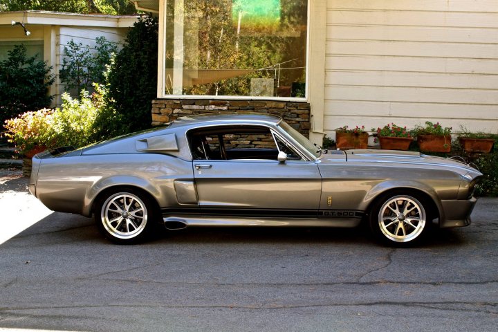 A black and white photo of a car parked in a parking lot - Pistonheads - The image displays a vintage Ford Mustang, which is a two-door, fastback coupe. The vehicle is silver and parked in front of a beige house with a brown brick wall. On the left side of the photo, greenery and a patio add a touch of nature to the scene. There are flowers visible in various pots and a small tree, indicating that the house has a well-maintained garden. The overall aesthetic of the image is one of nostalgic elegance, with the classic design of the Mustang juxtaposed against a modern residential setting.