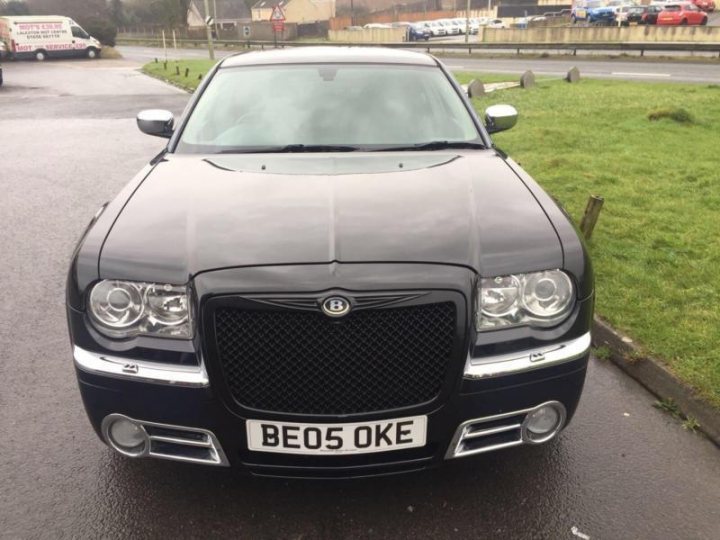 misbadging cars - Page 151 - General Gassing - PistonHeads - The image shows a black luxury car parked on a damp asphalt surface next to a grassy area. The car has a shiny, glossy finish and is equipped with front and rear fog lights, silver grille, and a navigation system. Its registration plate reads "BE05 OKE." The setting appears to be suburban or residential, with other vehicles and street signs visible in the background. The overall condition of the car and its shiny exterior suggest it's well-maintained.