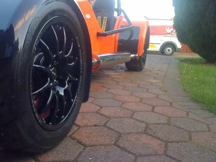 Pistonheads Contribution - The image features a close-up view of the front wheel of a unique orange jeep vehicle. The wheel is black and has a noticeable asymmetric design, with the thickness distancing slightly from the rim towards the front. The vehicle is parked on a brick driveway, indicating a residential setting. Behind the Jeep, portions of other vehicles can be glimpsed, suggesting that the Jeep is part of a group of parked vehicles. The sky appears to be cloudy, casting a diffused light over the scene.