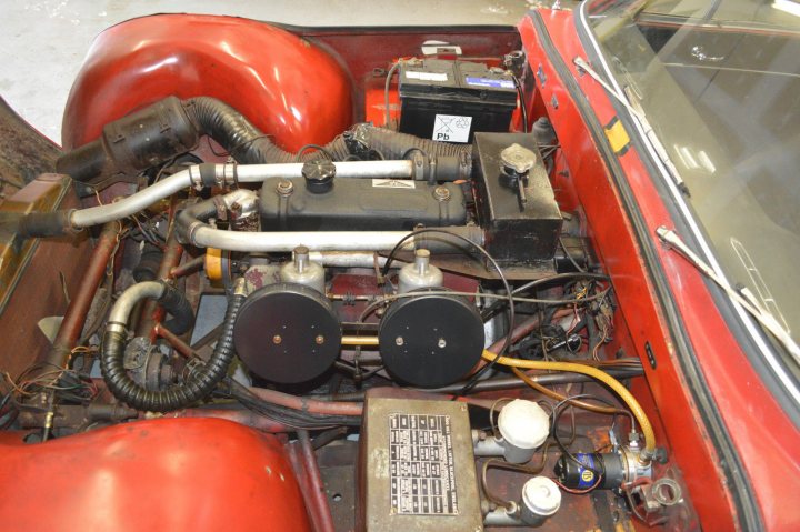 Early TVR Pictures - Page 90 - Classics - PistonHeads - The image shows an open hood on the rear side of a car. The engine is prominently visible, with various components and wiring visible. There is a large air filter on the right side of the engine bay. The bay is bathed in the light of an overhead fluorescent tube, and the paint of the trunk is visible, revealing a remnant of a past color change. A license plate is mounted on the trunk area. The style of the image suggests it might be a photograph from a mechanic's perspective or a stock photo used for educational or professional purposes.