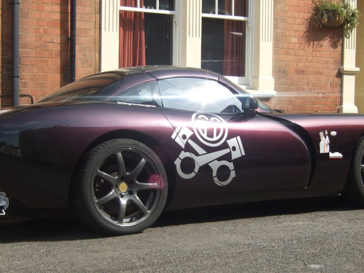 Your Car Stickered Up For Le Mans 2010! - Page 14 - Le Mans - PistonHeads - The image showcases a vintage sports car parked in front of a brick building. The car is a striking purple color with a black top, and it features sleek black wheels and a signature graffiti-style logo consisting of a circle surrounded by intricate patterns and a spanner on the hood. The building behind the car has a traditional red brick facade and is topped with a small garden or planter. A small plant can be seen in the planter, adding a touch of greenery to the scene. There's a paved sidewalk to the left of the car. The overall scene suggests an urban setting with a prominent display of car culture.