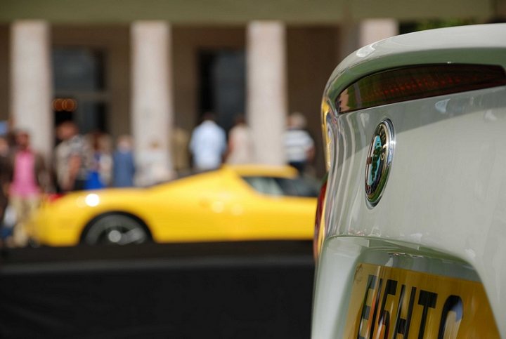 GRRC Lunch Club - Page 1 - Goodwood Events - PistonHeads - This image is a close-up photograph of a white car featuring a Licence Plate with the characters EL0TE. Behind the car, there is a blurred background with people and a yellow vehicle, which suggests a busy urban setting. The focus and composition of the image emphasize the car, with the background deliberately out of focus to draw attention to the car's details, license plate, and brand emblems. The overall image has a vibrant, slightly grainy texture, and the lighting suggests a bright, possibly sunny day.