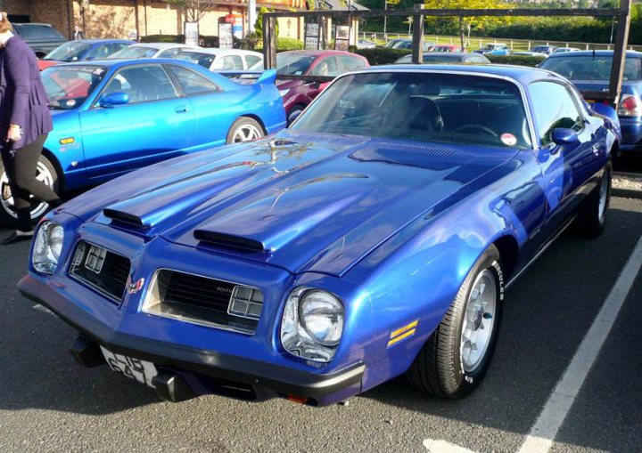 Pistonheads - This image captures a vibrant scene from a parking lot dominated by the deep royal blue camaro muscle car prominently positioned in the foreground. The camaro's sleek design and polished finish stand out against the various other vehicles scattered throughout the lot. A few individuals can be seen in the background, engaged in conversation near the vehicles. The setting appears to be during the day, as indicated by the natural light illuminating the scene. The image conveys a sense of movement and activity, with the camaro being the focal point of attention.