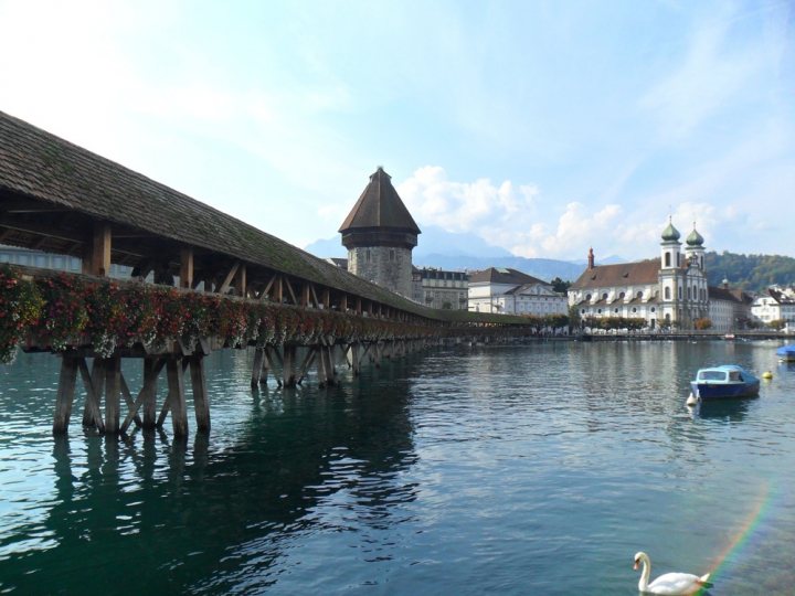 Back from Europe - Page 1 - Aston Martin - PistonHeads - The image showcases a picturesque lakeside village with a traditional wooden barn alongside a calm body of water. At the end of the pier, there's a charming Swiss-style house with a gabled roof, emphasizing a quaint and tranquil atmosphere. Panoramic views of the lake surround the pier, leading to a distant glimpse of majestic mountains. Above, the sky is a beautiful canvas of blue, dotted with fluffy white clouds. On this idyllic scenery, a lone swan gracefully glides through the water, adding a touch of serene beauty to the scene.