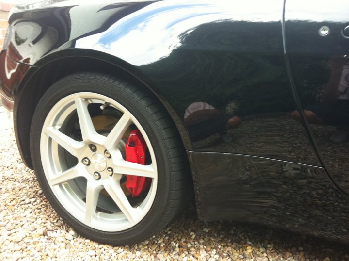 Rear 1/4 Protective Film - Looking grubby - Page 1 - Aston Martin - PistonHeads - This is a photograph featuring a close-up view of a car's rear tire. The tire is split into two different rims, with one half being white and the other red. The layers of the tire's structure are visible due to a reflection in the image, enhancing the contrast and highlighting the tire's inner details. The car's metallic bodywork glistens, reflecting an array of colors due to the light. The background appears to be a textured surface, possibly a gravel driveway or parking area, and it offers a contrasting backdrop that emphasizes the car's shiny appearance.