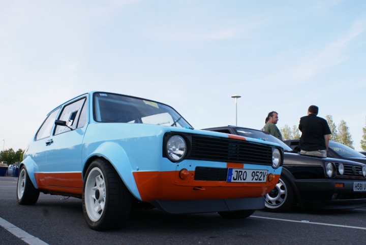 Pistonheads Present - This image captures a vibrant scene on a street corner. The main focus is a distinctive blue-orange car parked on the side of the road. The car is old and appears to be well-maintained. It has a large front grille and a spoiler mounted on the top. The license plate of the car is clearly visible. In the background, we can see two more cars parked further down the street, as well as a solitary lamppost. There are also three people in the vicinity of the blue-orange car, but their actions are unclear from this angle. The overall atmosphere of the image is casual and relaxed, with the bright colors of the car standing out against the more muted tones of the surrounding environment.