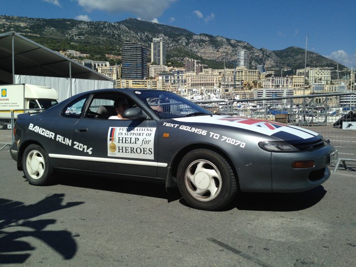 7 Countries 4 days & a banger - 'Help For Heroes' - Page 1 - South West - PistonHeads - This image features a striking black car prominently displayed in the foreground. The car is adorned with a bumper sticker in the shape of a banner. The banner proudly declares "Run 2014" in bold white letters on a black background, indicating a possible event or cause related to running. 

A noticeable feature on the car is a license plate that reads "B augmenting". A small British flag is also seen affixed to the rearview mirror, suggesting a sense of pride or association with the United Kingdom. The backdrop of the image reveals a cityscape dotted with buildings and mountains, adding depth to the scene. 

The car appears to be stationary, possibly parked on the street. The presence of the flag suggests that it might be used for driving or advertising within the city. The overall composition of the image places the car as the central focus, with the cityscape subtly enhancing the setting.