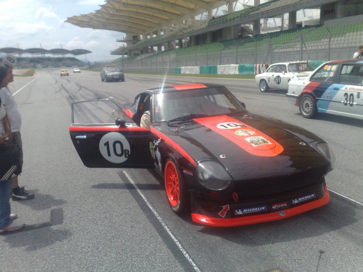 Pistonheads Afos Sepang - In the image, there is a striking black racing car prominently displayed in the foreground. This car appears to be a tribute to the iconic 'Batmobile', complete with a red, white, and black body and the number 10 emblazoned on its side. It's parked on a racetrack, surrounded by a sense of speed and excitement that's palpable even in the stillness of the image.

The slick, black top of what appears to be a luxury sports car is visible in the background, adding a touch of elegance to the otherwise gritty racetrack scene. The railing and bleachers above the track suggest that this is a professional racing venue, designed for high-speed action.

Aside from the cars, there are also several people present in the image. They are scattered along the sidelines, likely spectators or crew members of the competing cars. Their exact actions and interactions are not clear, but their presence adds an intriguing human element to the scene.

Overall, the image captures a moment of quiet before a flurry of action, with the shiny black racing car waiting for the starting gun. The atmosphere is tense but