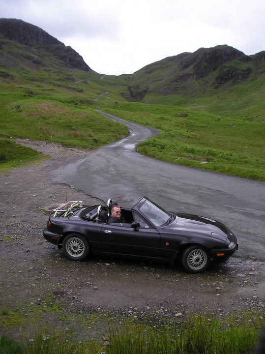 One NA, two holidays, 3000 miles - one change of  heart... - Page 1 - Mazda MX5/Eunos/Miata - PistonHeads - In the image, there is a man sitting in an open black sports car, which is parked on a muddy gravel surface. The backdrop features a picturesque green field dotted with scattered rocks. A winding, uphill dirt road radiates into the distance, bordered by grassy embankments and a rocky crag. The sky above is filled with gray, overcast clouds, casting a muted light over the entire scene.