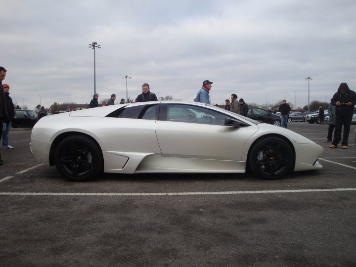 Sunday Service Pistonheads Oxford - The image depicts a sleek, low-profile sports car in a parking lot. The car, which has a futuristic design with gull-wing doors, is white with black accents. It's parked diagonally across multiple parking spaces, taking up a significant portion of the view. In the background, there are a number of people walking around, and other cars parked in the lot. The sky is overcast, creating a cool, grey tone to the image.