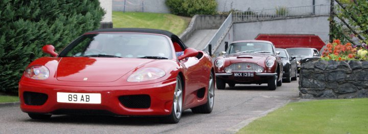 Astons & Ferraris at Glenfiddich distillery - Page 1 - Aston Martin - PistonHeads - The image captures a scene of vintage cars moving down a narrow road bordered by a grassy area and stone walls. Two of the cars, a prominent red convertible and a similar vintage model further back on the road, stand out against the stone walls and lush grass. The setting is outdoors and most likely in an urban or residential area, given the road and vehicles. The style of the cars suggests that this could be a special event or a themed gathering dedicated to vintage and sports cars.