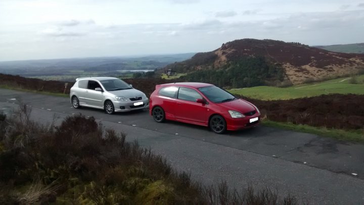 Civic EP3, BMW 130i, Westfield XTR2 - Page 1 - Readers' Cars - PistonHeads - The image captures a picturesque scene of a mountainous landscape with two small cars parked on the side of a road. A white car and a red sports car are positioned side by side, parked adjacent to the road. The cars are facing the same direction, suggesting they have just stopped for a brief moment. The road leads to a scenic overlook, offering a breathtaking view of the surroundings, with a mix of grassy fields, brown hills, and a body of water that adds to the tranquil setting. The overall mood of the image is serene and peaceful.