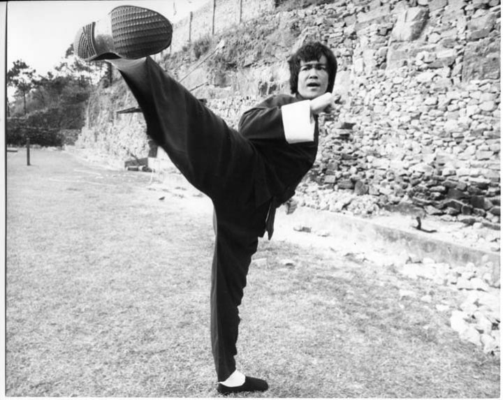 In this vintage black-and-white photograph, a martial arts practitioner is frozen in a powerful and balanced kick. The individual, dressed in a traditional martial arts outfit and shoes, is gripping a device near the bottom of the frame, possibly a weapon or a training aid. The setting appears to be a dilapidated outdoor area, with walls and rocks forming a rough background. The stance and expression of the person suggest focus and mastery of their art.