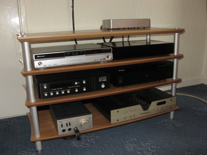 Hifi Set Pistonheads - The image displays a well-organized wooden shelf against a patterned carpeted wall. On the shelf, there is an array of electronic equipment, including what appears to be stereo and other home entertainment components. The devices are neatly arranged in two rows, with one row on the top shelf and the other on the bottom. The top shelf is slightly short of its counterpart, leaving a small gap. The shelf has a metal frame, enhancing its sturdiness. The entire setup looks like a home entertainment center.