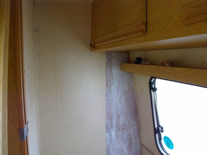 My caravan history - Page 1 - Tents, Caravans & Motorhomes - PistonHeads - The image depicts a compact, well-organized interior space. A small cabinet is mounted on a beige wall, with brown wooden doors. The cabinet has two thick shelves, enabling storage of items, and a thin horizontal space at the bottom, possibly for hardware or tools. In the cabinets, there are several items, but their exact nature is not clear from the image. A window with a partially visible blue circle is situated next to the cabinet on the front right side. The window lets in light, and the overall design suggests efficient use of space, likely in a boat or a similarly confined area.