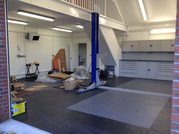 Who has the best Garage on Pistonheads???? - Page 193 - General Gassing - PistonHeads - The image depicts a storage room in the process of renovation. The room features open shelves, a ramp, and various tools and materials scattered around. Wooden crates are visible in the lower right corner, and a large sheet of drywall is mounted on the floor. There is natural light coming in from the upper left corner where the room connects to another room.