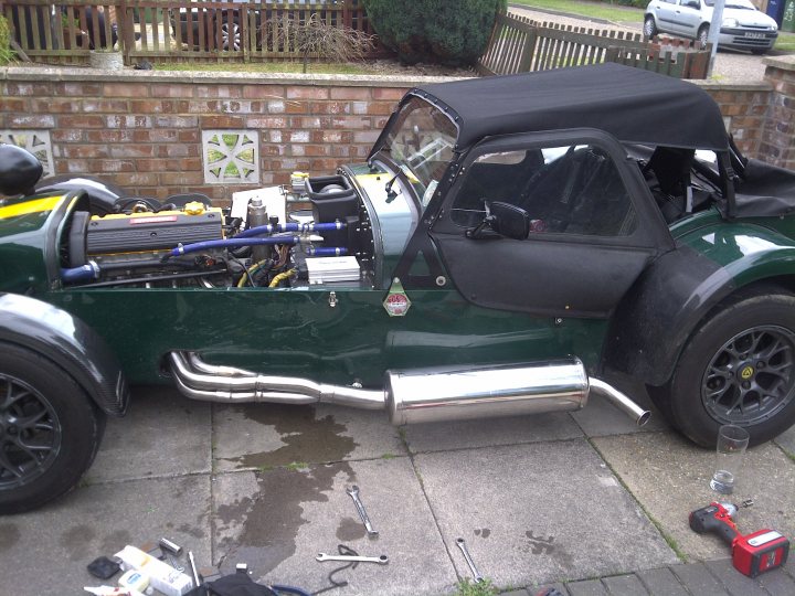 Repacking a sealed exhaust?? - Page 1 - Caterham - PistonHeads - The image shows a vintage car parked on a driveway or sidewalk. The car's rear end is visible and appears to have a spare tire mounted on the back. It has a distinctive military green color scheme. There are visible remnants of what looks like various car parts on the ground around the car, indicative of recent mechanical work or even a DIY repair job. There is a ladder at the entrance of the vehicle, suggesting it may have been used for access or storage purposes. Additionally, there's a cup of water next to the car, which could be for hydration during the work or to clean components.