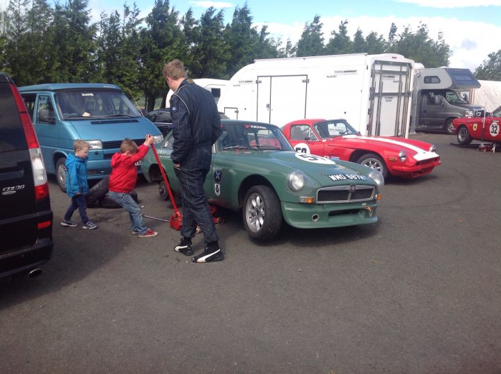 Knockhill Paddock Pics - Page 1 - UK Club Motorsport - PistonHeads - The image captures the scene of an outdoor motorsport event, featuring a man leaning on a bright green vintage car with the model "MGB" visible on the side, and two young boys, one in the middle holding onto an orange pole, with another one on the far right. The car shares some space with a red sports car to the right, and other vintage cars are visible in the background. There's a white camper to the left and a gray trailer on the far right, suggesting a spacious venue for this event.