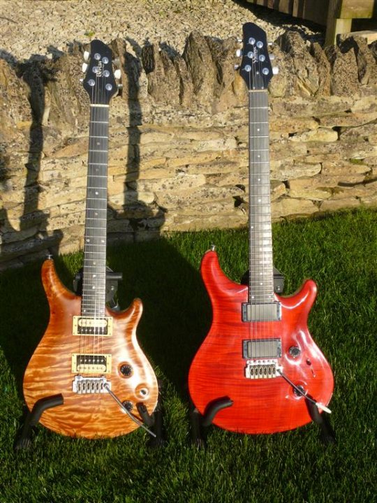 Pistonheads - The image displays two guitars, one cherry wood-finished and the other a solid-body red guitar, resting on grass. Both guitars are placed upright, leaning against a rock or stone wall with weathered textures. The solid-body red guitar is positioned on the right with cords attached to its upper bout.