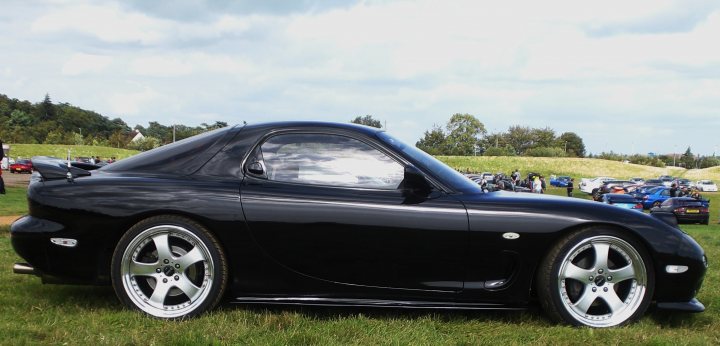 Results Pistonheads Mapped Rex - This image features a sleek, black convertible car parked on a grassy field. The car is shiny and characterized by its numerous air vents along the hood. It has a black hardtop roof, which is currently closed, suggesting that the photo may have been taken after or before a ride, perhaps at an outdoor gathering. Behind the car, the backdrop consists of a clear sky divided by a few clouds, adding a serene background to the scene. Additionally, there's a line of cars parked in the distance, indicating that this field might be a parking or display area for vehicles.