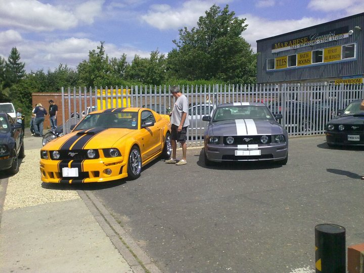 July Sports Pistonheads Atlantic Sat Bbq - The image captures a scene in a parking lot, with a man standing between two cars. The man is wearing a white shirt and black shorts, and he is facing away from the camera, possibly admiring or examining the cars. 

The car closest to the man is a yellow Mustang with a black stripe on the hood and a black stripe on the top. It has white and black stripes on the sides as well. The other car, further away, is a silver car with a black stripe. The parking lot is surrounded by a metal fence, and there are a few other cars and individuals visible in the background. The setting suggests a casual, outdoor gathering or event, possibly related to the Mustang car.